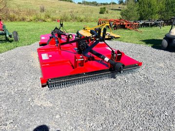 Red rotary cutter on gravel with farm equipment in the background.