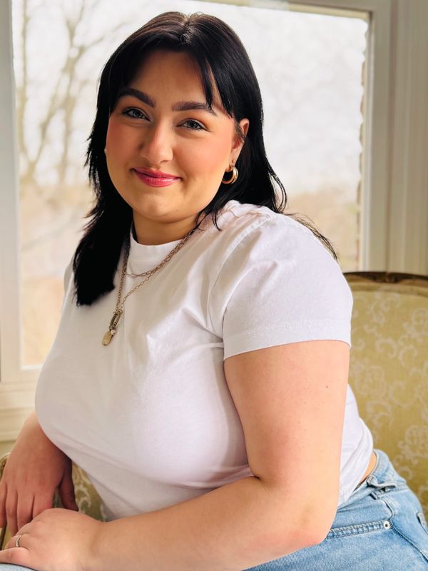 Smiling woman in white t-shirt sitting by a window with natural light.