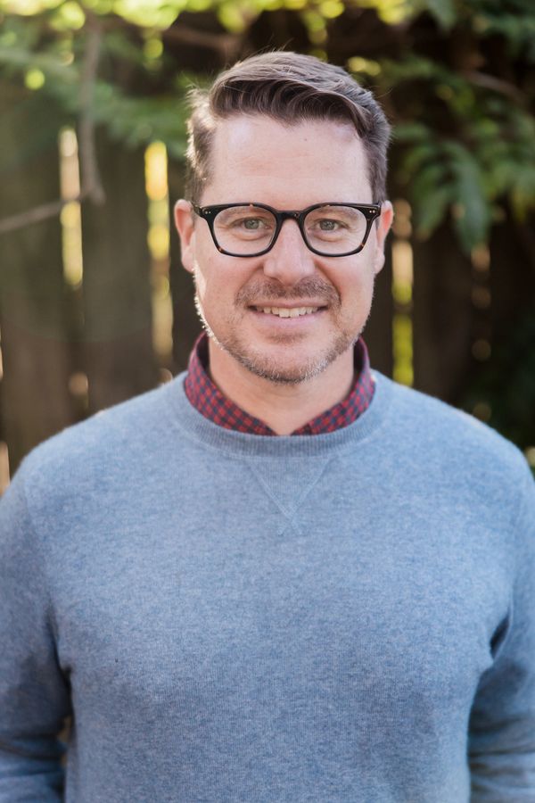 Smiling man with glasses in a blue sweater outdoors in natural light.