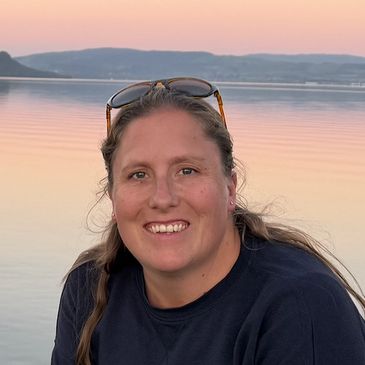 Smiling woman with sunglasses on her head by a serene lake at sunset.