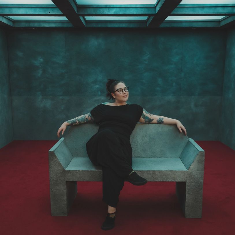 Woman with tattoos and glasses relaxes on a modern concrete bench in a minimalist room.