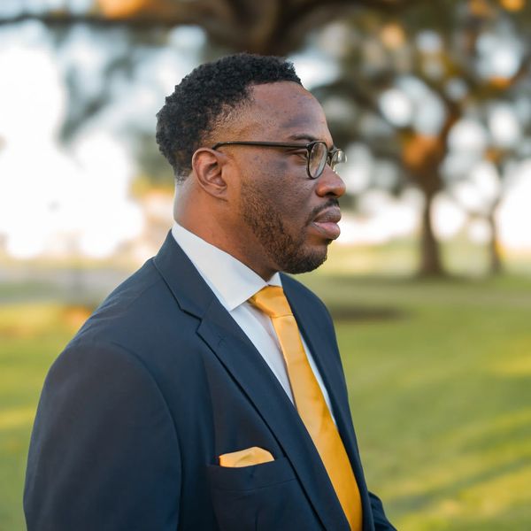 Man in a suit with yellow tie standing outdoors in profile.