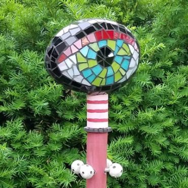 Colorful mosaic eye sculpture on a pole in greenery.