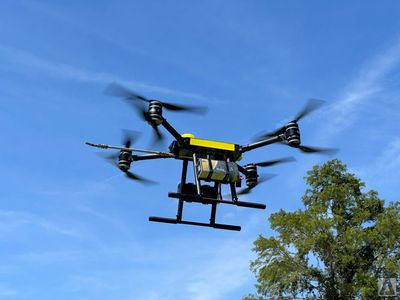 A picture of a CleanFlight Solutions drone flying in the clear blue sky withtrees in the background.