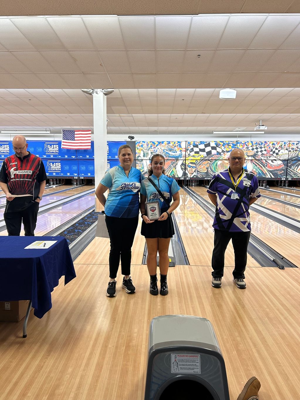 Three people standing on a bowling lane with one holding an award plaque.