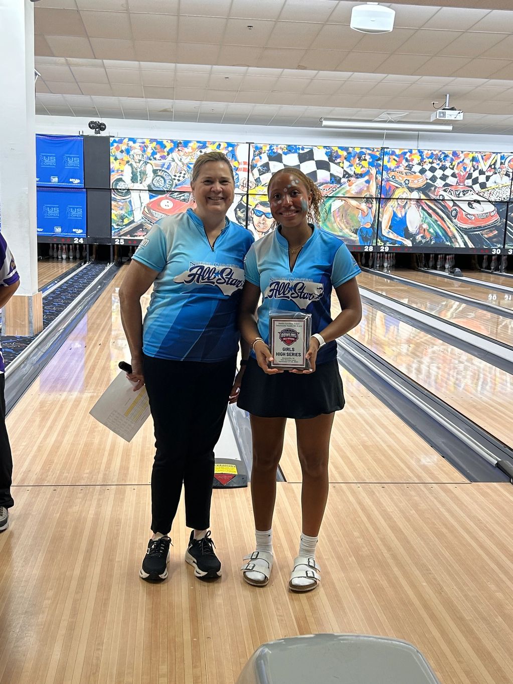 Two smiling women in bowling uniforms at a bowling alley, one holding a trophy.