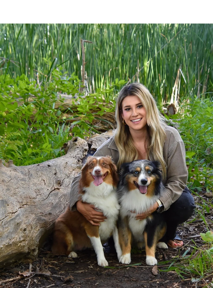 Woman happily poses with two Australian Shepherd dogs in a lush outdoor setting.