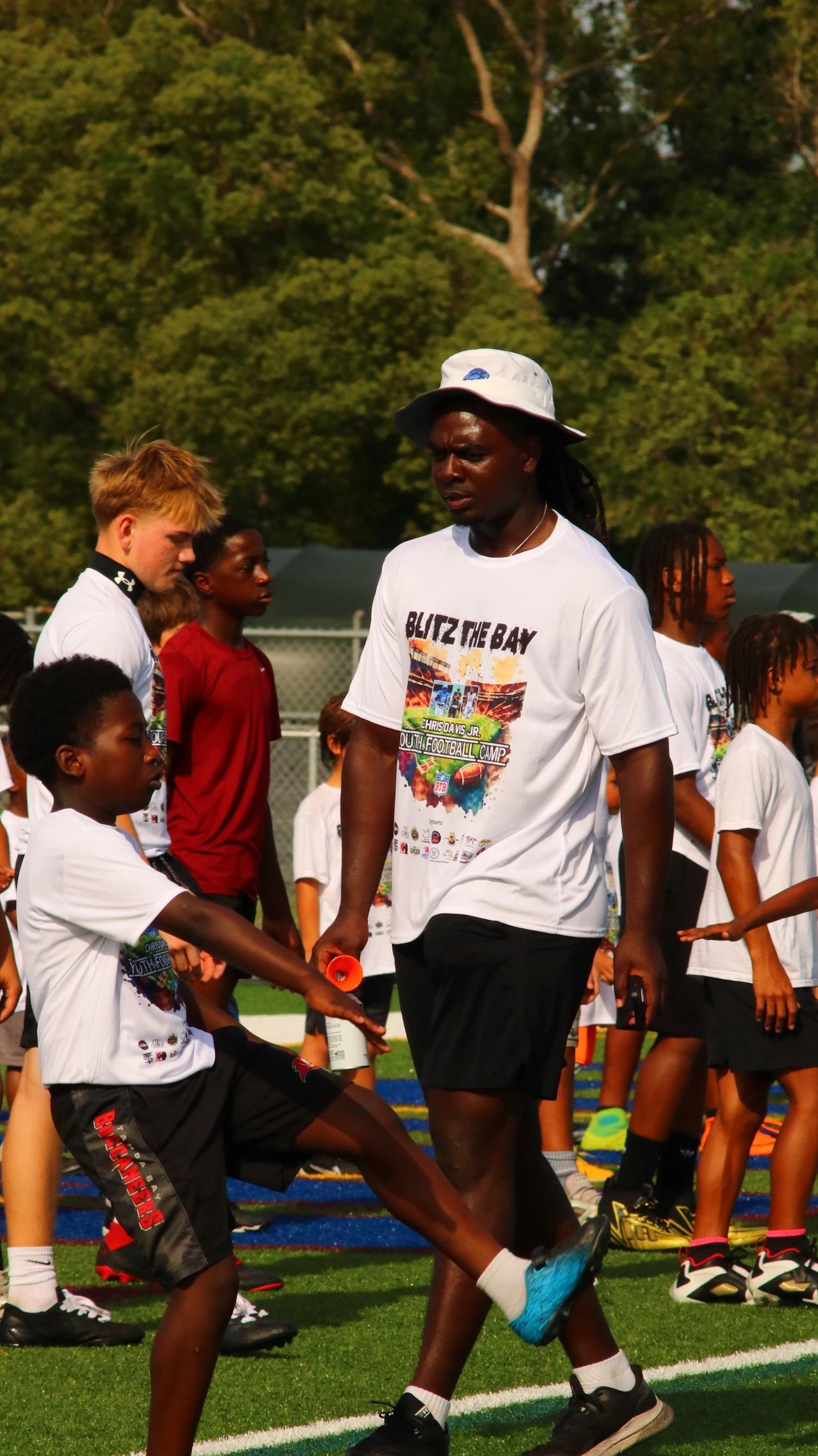 Coach supervises kids stretching at a youth football camp on a sunny day.