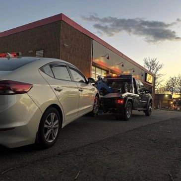 Tow truck towing a white car at dusk near a building.