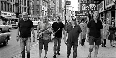 A group of six men walking on a busy city street in black and white.