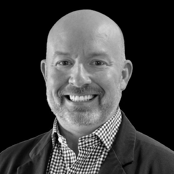 Smiling bald man with a beard in a checkered shirt and dark jacket against a black background.