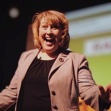 Woman enthusiastically speaking on stage with a bright smile.