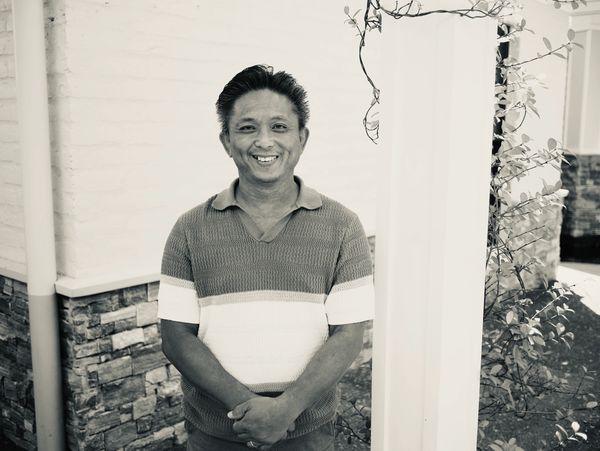 Black and white portrait of a smiling man standing outdoors near a pillar.