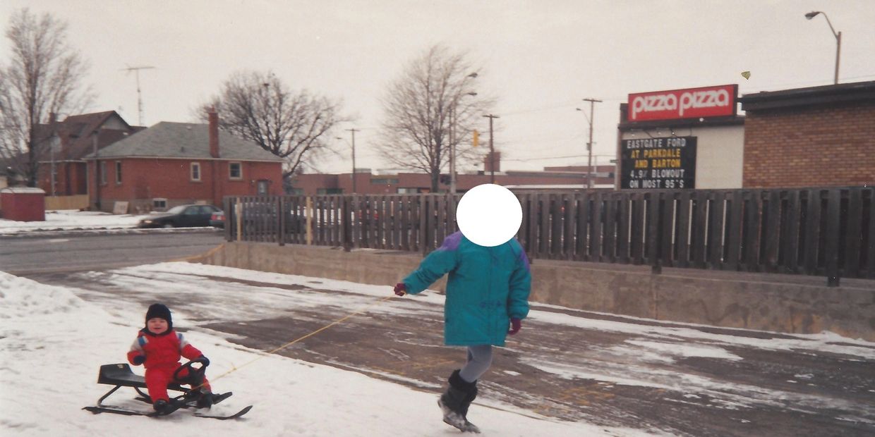 Child pulls a toddler on a sled in a snowy urban area.