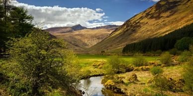 Scenic valley with a stream flowing through lush greenery and hills under a partly cloudy sky.