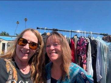 Two women smiling outdoors with clothes hanging behind them.