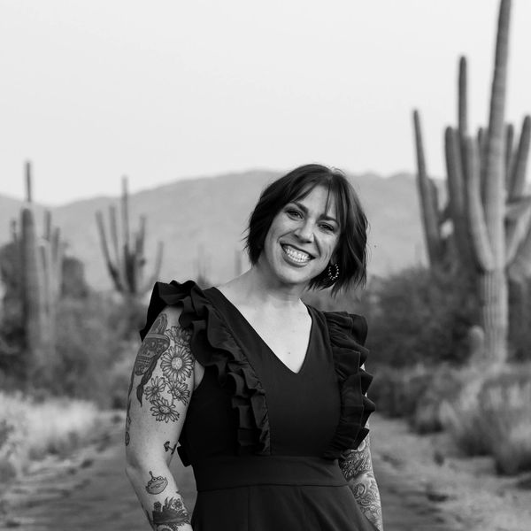 Smiling woman with tattoos stands on a desert path surrounded by cacti.