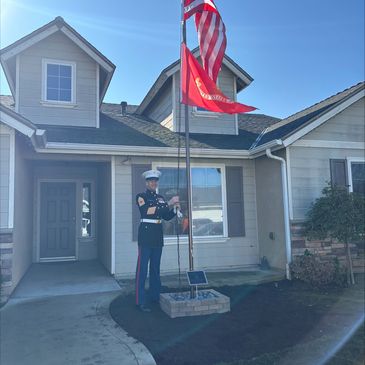 U.S. Marine raising the American and Marine flags outside a home on a sunny day.