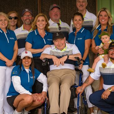 Group photo of diverse adults and a baby, with a man in a wheelchair, all smiling in matching polo shirts.