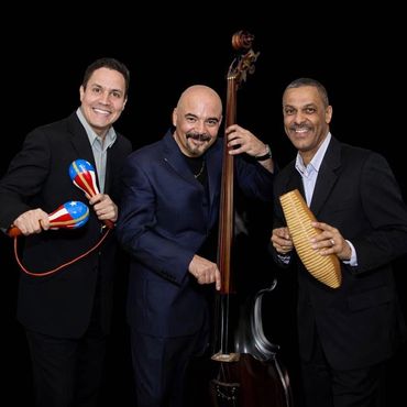 Three musicians posing with percussion and string instruments against a black background.
