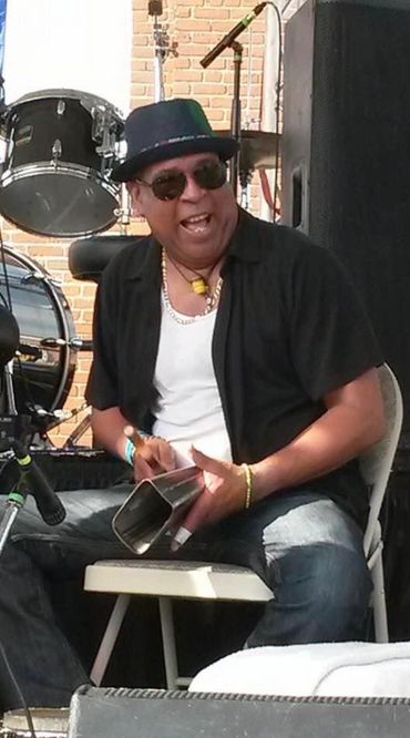 Man playing a cowbell with a smile, wearing sunglasses and a hat.