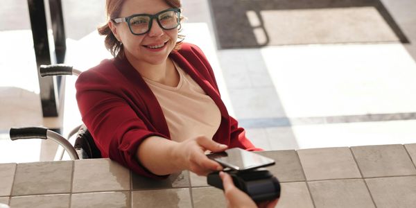 A young woman in a wheelchair reaches over a counter with her phone to pay electronically.