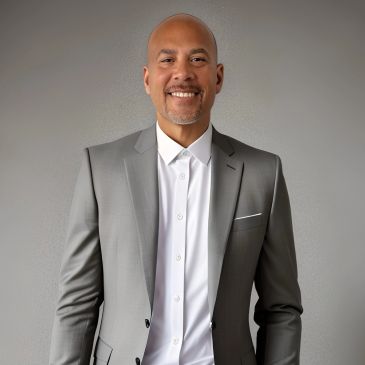 Smiling man in a grey suit and white shirt standing against a plain wall.