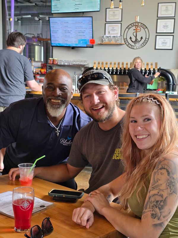 Three friends smiling at a bar with drinks and a tattooed woman.
