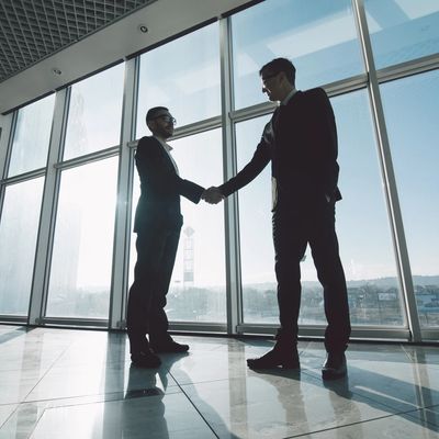 Two young businessmen shake hands with each other standing against panoramic windows.
