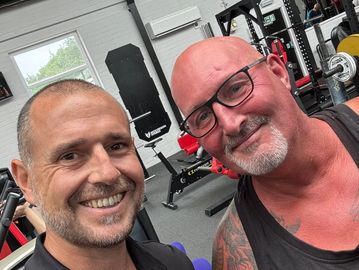 Two men smiling inside a gym with workout equipment in the background.
