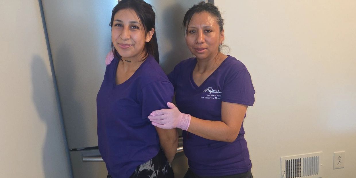 Two women in purple shirts, one wearing pink gloves, posing indoors against a plain wall.