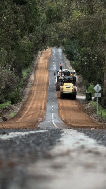 Road construction with heavy machinery on a hilly, tree-lined road.
