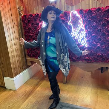 Woman posing stylishly in cowboy hat and boots by a neon angel wings sign.
