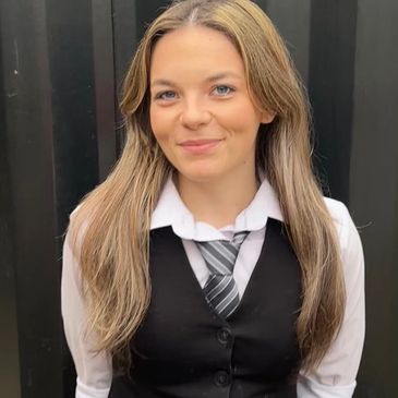 Young woman smiling, wearing a black vest and tie against a dark background.
