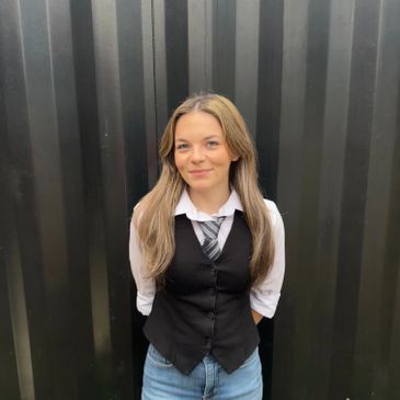 Young woman smiling, wearing a black vest and tie against a dark background.