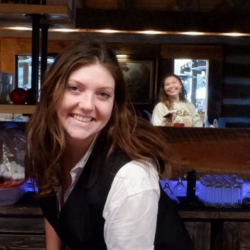 Smiling bartender posing behind a rustic wooden bar in a cozy setting.