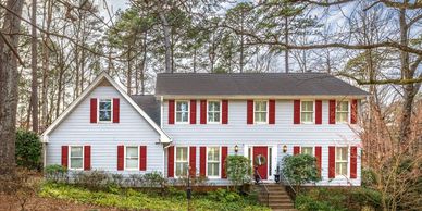 Two-story white house with red shutters and front door, surrounded by trees.