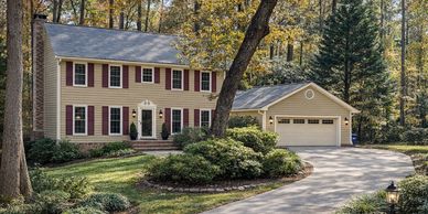Two-story suburban house with beige siding and red shutters surrounded by trees.