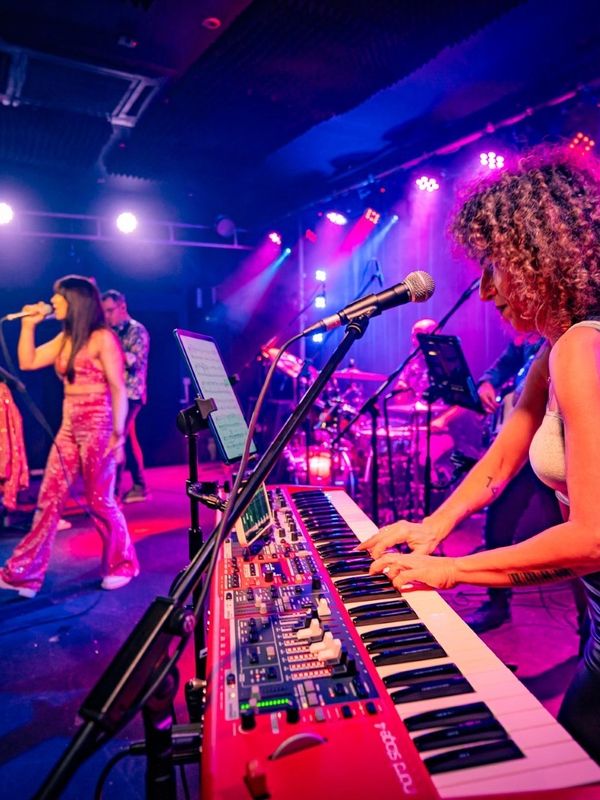 Woman playing keyboard on stage with band and singer under colorful lights.