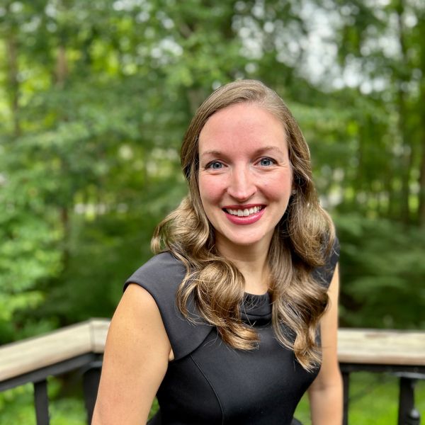 Smiling woman with wavy hair in a black dress standing outdoors on a deck.