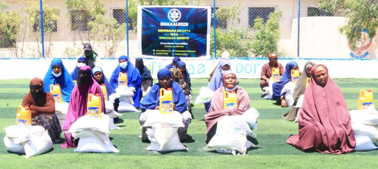 Group of women sitting with food aid packages on green field under a banner.
