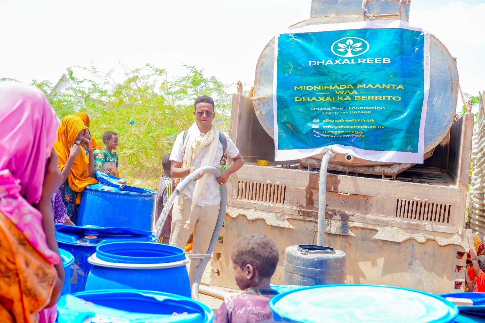 A man distributes water to people using blue containers from a DhaxalReeb truck.