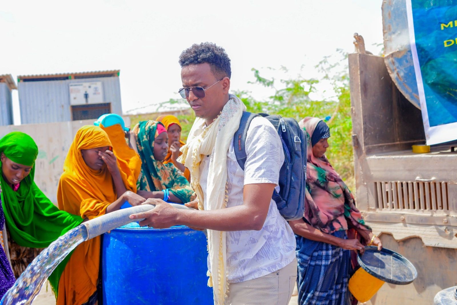 People collecting water from a hose into containers in a sunny outdoor setting.