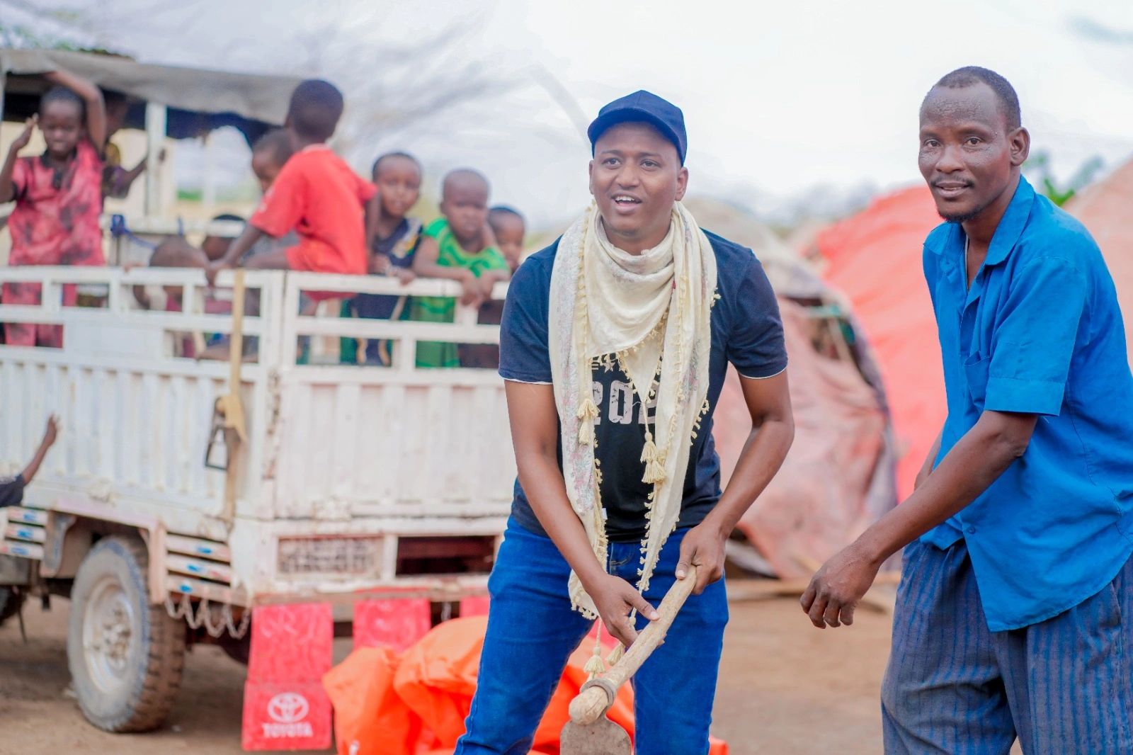 Two men holding a farming tool with children watching from a truck in the background.