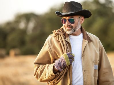A bearded middle aged man in a cowboy hat and leather jacket with a lasso over his right shoulder