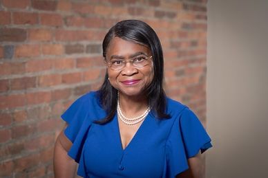 Professional woman in a blue dress with glasses and pearls, smiling confidently.