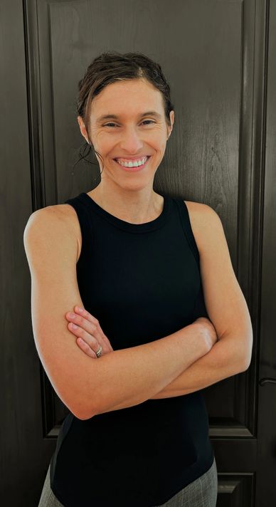 Smiling woman in a black sleeveless top stands with arms crossed against a dark wooden background.