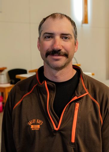 Man with a mustache wearing a brown jacket indoors.