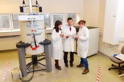 Three scientists in lab coats discussing results near a large scientific instrument.