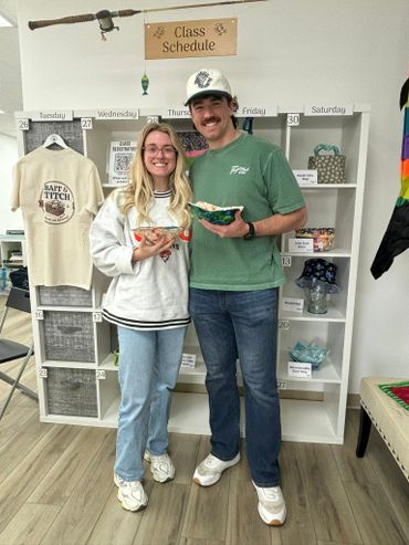 Two smiling people holding bowls in a cozy room with shelves and a class schedule.
